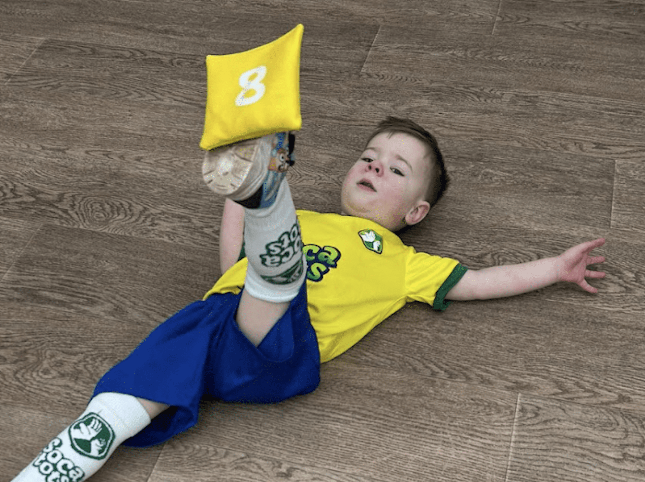 Image shows a Socatots child balancing a bean bag on his foot.